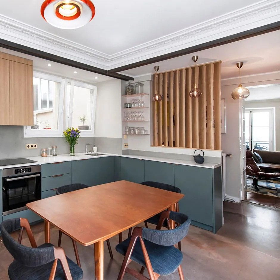 Vue sur une cuisine ouverte avec meubles bleu vert et aménagements bois, claustra de séparation et table de salle à manger